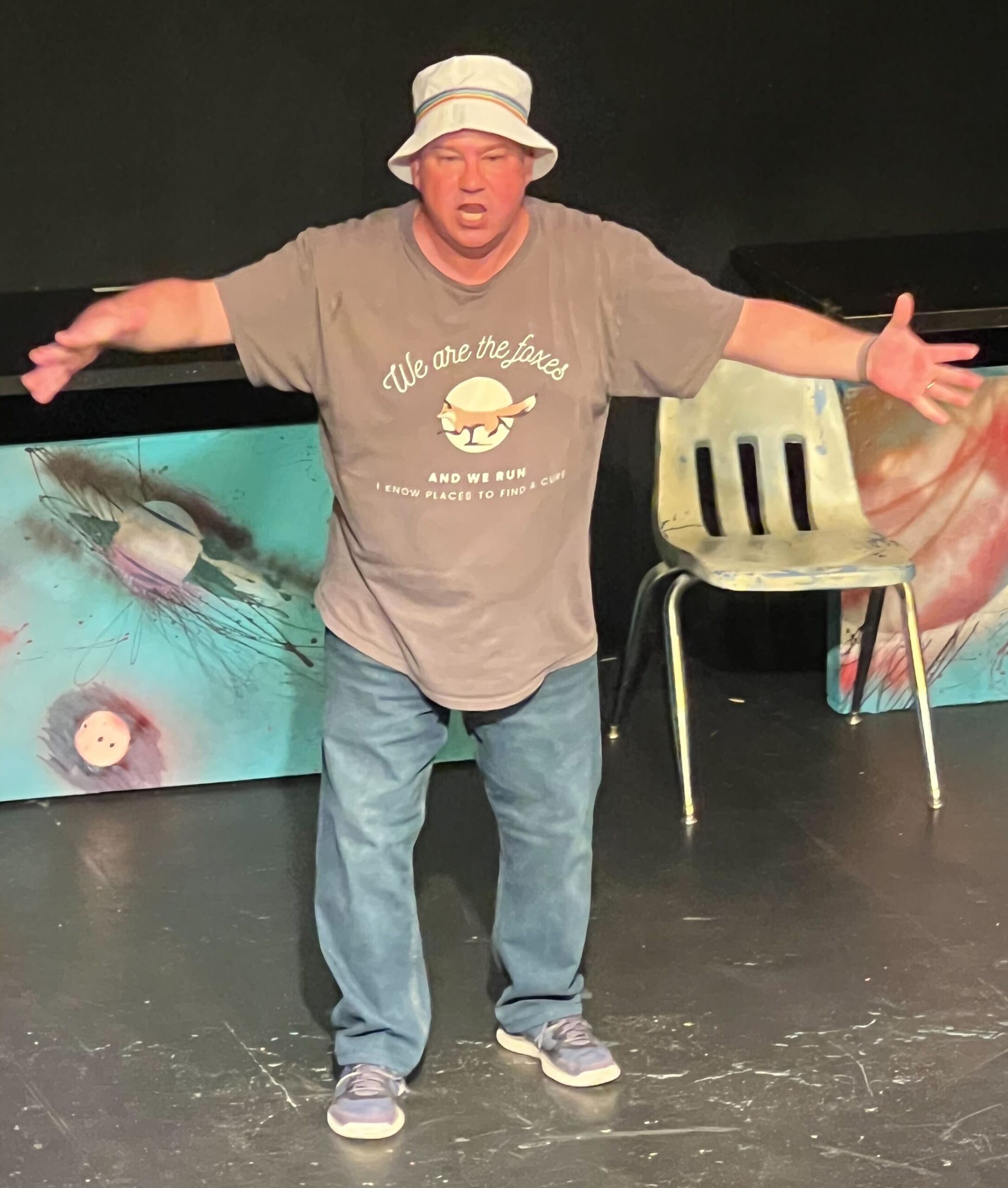 A man wearing a bucket hat, grey t-shirt and jeans gesturing with his arms wide open